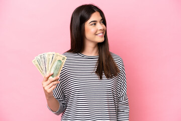 Young Brazilian woman taking a lot of money isolated on pink background looking to the side and smiling