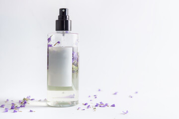 Bottle with natural floral water spray and purple flowers petals stands on white background.