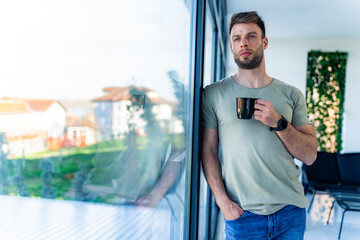 A man is drinking his morning coffee leaning against the window, his hand is in his pocket and he...