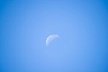 Wieczorne, bezchmurne, niebieskie niebo. W środku kadru widać białą tarczę księżyca. Księżyc wchodzi w fazę ostatniej kwadry.