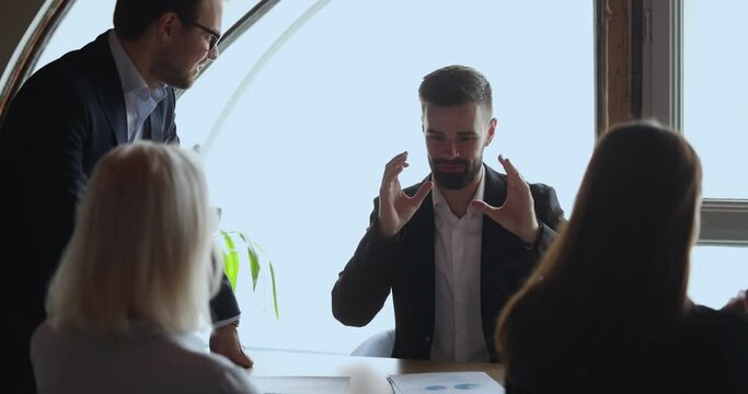 Cheerful handsome young employee telling funny story on meeting, talking to colleagues, speaking, laughing, sitting at conference table. Happy coworkers discussing creative ideas, enjoying teamwork