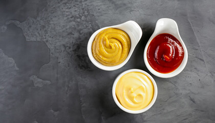 Obraz na płótnie Canvas Set of three sauces - mayonnaise, mustard and ketchup in white ceramic bowls on black stone or concrete background. Selective focus. Top view.