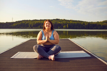 Young woman gets to unwind and refill with energy in nature. Relaxed fat chubby woman sitting in yoga position on wooden pier on calm sunny summer morning, with beautiful lake or river in background