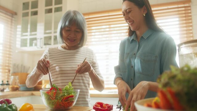 Happy attractive Asian elderly woman mom and daughter enjoy cooking together mixing salad in bowl pick vegetable on wooden cutting board preparing ingredient of healthy vegetarian food in cozy kitchen