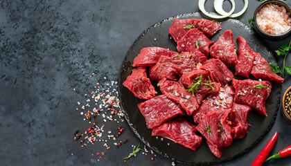 Cut beef into small pieces with sea salt, dried herbs and chili peppers on dark slate or concrete background. Top view.