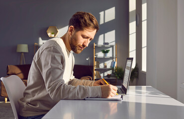Side view of a young attractive business man working on a laptop at the office or at home and writing at the desk making notes, calculations, considering new projects, analyzing company.