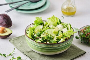 Healthy food, green salad with avocado, cucumber, microgreens and feta cheese in a green salad bowl on a light concrete background. Green monotone.