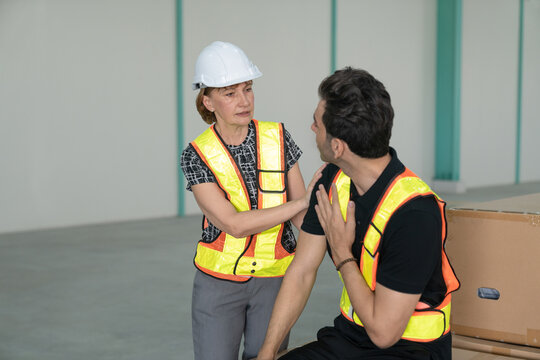 Woman factory engineer comforts a strained male engineer inside a large industrial warehouse.Male factory engineer with shoulder injury sitting on cargo box with female engineer helping.