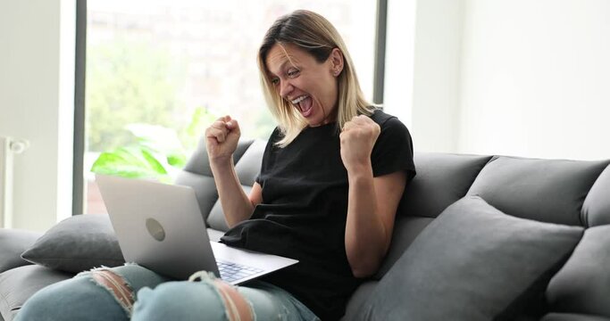 Woman is sitting on couch, clenching fists and screaming with joy and reading great news on laptop. Girl celebrating victory in online auction