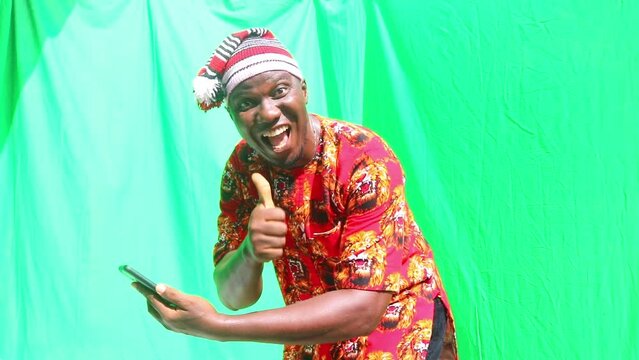 Nigerian Igbo Business man in local red dress displays excitedly at news on his phone and happily approves with a thumb up