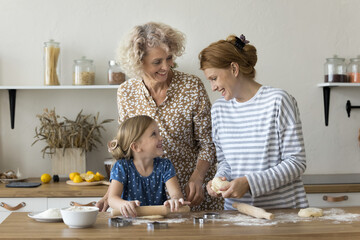 Happy adorable kid, positive mom and grandma baking in home kitchen together, rolling dough on table, preparing homemade bakery food, dessert, cookies, talking, laughing