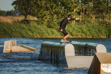 Beautiful view of young man rider holding rope and making extreme jump on wakeboard. Wakeboarding and water sports activity. Low angle shot of man wakeboarding on a lake. Man water skiing at sunset.