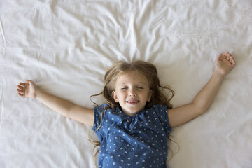 Happy sleepy preschool child girl lying on white sheet on bed with closed eyes, open arms, toothy...