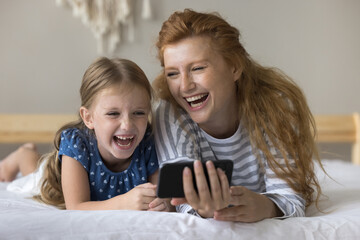 Cheerful pretty red haired mom showing content smartphone to cute daughter girl. Happy mum and little kid relaxing at home, using online app, service on mobile phone, laughing