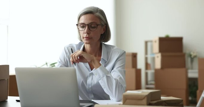 Thoughtful mature entrepreneur woman working at laptop in e-commerce storage room with stacked boxes, looking away, thinking over answer to customer, typing on computer