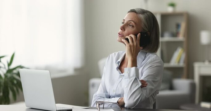 Successful mature freelancer woman working at home, using gadgets for communication, talking to customer on mobile phone at table with laptop, getting happy, smiling, laughing