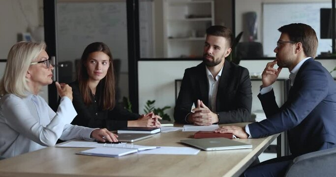 Serious older mature business woman speaking to listening, nodding colleagues at office meeting table, offering decision, idea for project, telling strategy. Diverse team discussing tasks