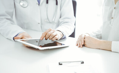 Doctor and patient sitting near each other at the desk in clinic. The focus is on female physician's hands pointing into tablet computer touchpad, close up. Medicine concept