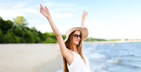 Fototapeta na wymiar Happy smiling woman in free happiness bliss on ocean beach standing with a hat, sunglasses, and rasing hands. Portrait of a multicultural female model in white summer dress enjoying nature during