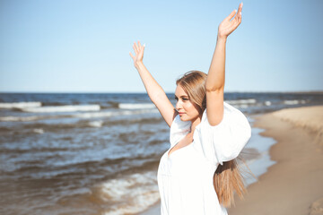 Fototapeta na wymiar Happy, beautiful woman on the ocean beach standing in a white summer dress, raising hands