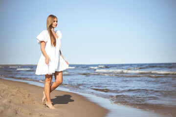 Happy smiling beautiful woman is walking on the ocean beach in a white summer dress