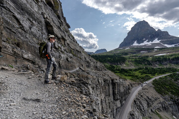 Matured Caucasian man, hiker, standing on a cliff watching a mountain scene while vehicles driving along a road below, Highline Trail, Going to the Sun Road, Glacier National Park, Montana