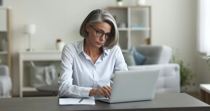 Serious middle aged business freelancer woman in glasses working at laptop at home, typing, looking at screen, touching chin, thinking over Internet communication, watching online content