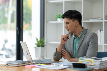 Young Asian businessman working with computer laptop in Analyze plans, search for document information, messaging keyboards, e-commerce, finance technology startup business ideas.