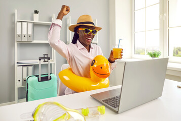 Funny happy smiling african american woman sitting on the desk on workplace at office with juice coctail in a beach rubber ring and booking tickets for summer vacation online via laptop.