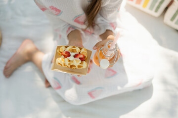 Top view of a little girl in a dress sitting on a blanket and holding strawberry waffles and juice in her hand