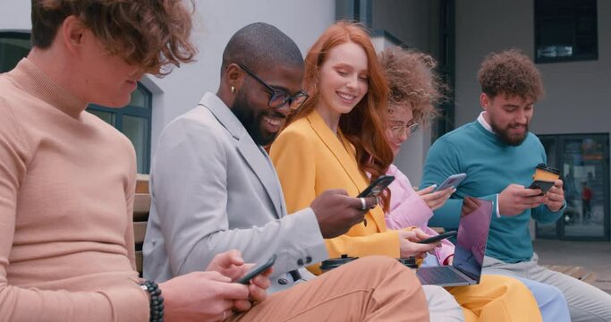 Group of business people in stylish colorful clothes are looking into their smartphones while sitting outdoors. Breaking time. Waiting for a meeting. Slow motion
