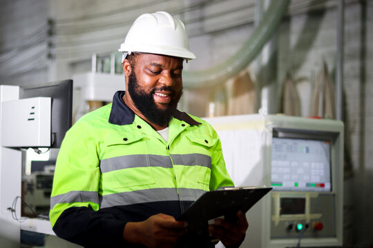 Industrial Worker Man With Whiskers Wearing Helmet And Safety Vest, African Engineer Holding File Folder Document At CNC Woodworking Carpentry Furniture Factory With Computer Controller As Background.