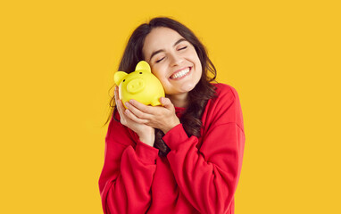 Portrait of happy smiling brunette woman snuggling up to yellow piggy bank on yellow background with closed eyes. She is happy about her savings. Saving money, self finance, savings, capital concept. - Powered by Adobe