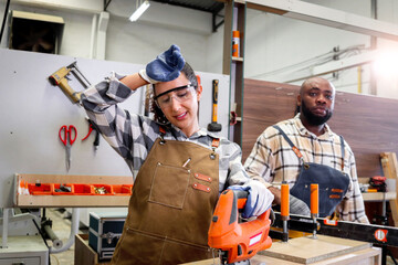 Exhausted beautiful carpenter woman wears safety glasses goggles and apron, works with coworker friend African craftsman at woodworking carpentry furniture workshop, female craft worker feeling tired. - obrazy, fototapety, plakaty