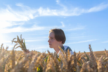 A young guy, a teenager with ears of ripe wheat in a farmer's field, against a blue sky. Grain for...