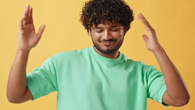 Video of a cheerful handsome Indian man with curly hair in a turquoise t-shirt dancing in a studio on a yellow background and gesturing with his hands. Relaxation and leisure.