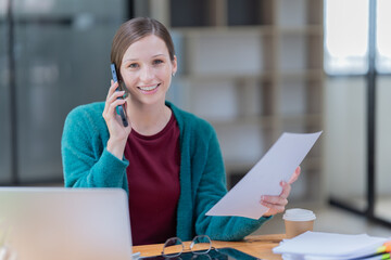 Sharing good business news. Attractive young businesswoman talking on the mobile phone and smiling while sitting at her working place in office and looking at laptop PC