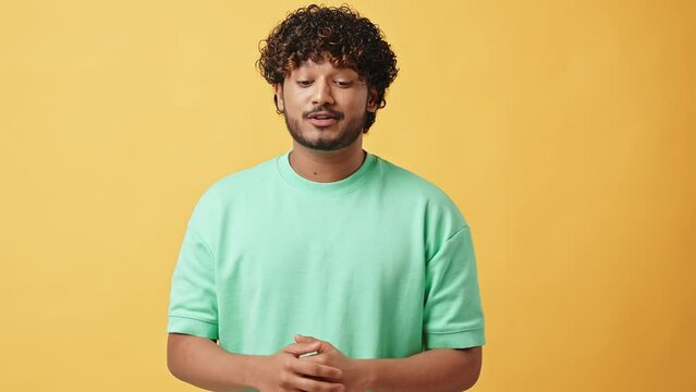 Handsome Indian man standing in studio on yellow background and talking to camera, pointing finger up, finger down to subscribe to subscriber channel, etc. The camera moves towards him with a slider.