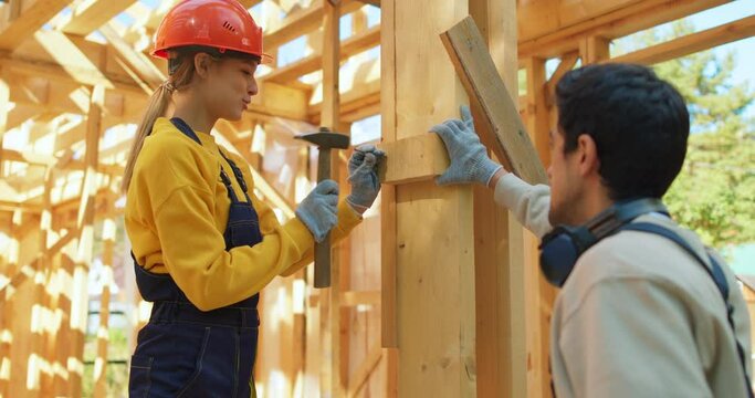 Caucasian young female engineer architect foreman labor worker wears safety gloves hardhat uniform holding using steel hammer pounding nail on stick Professional male carpenter helping