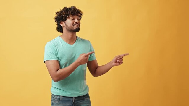 Advertising video portrait of a handsome curly-haired, young, positive, stylish Indian man who stands on a yellow background and points his fingers to the side at an empty place and calls to look here