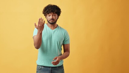 Advertising video portrait of a handsome curly-haired, young, positive, stylish Indian man standing on a yellow background invites you to come here. Copy space.