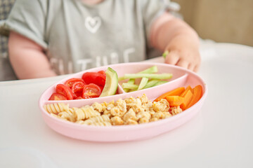 Obraz na płótnie Canvas Cute child eats healthy food pasta and vegetables steamed,. Portraits of a cute 10 months old baby girl. The baby sitting in a special high chair for babies. 