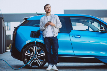 Young man charging electric car at home and using mobile phone