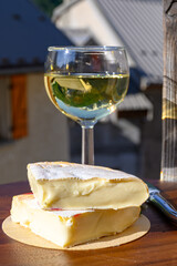 Reblochon French cheese made in Haute-Savoie, white Roussette de Savoie or Vin de Savoie wine with view on old alripne village and Hautes Alpes mountains snow tops in summer
