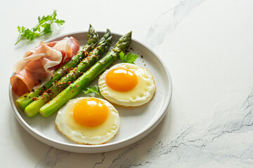 Breakfast fried eggs with fresh asparagus and ham (jamon), herbs in a plate on a white background. Delicious hearty brunch.