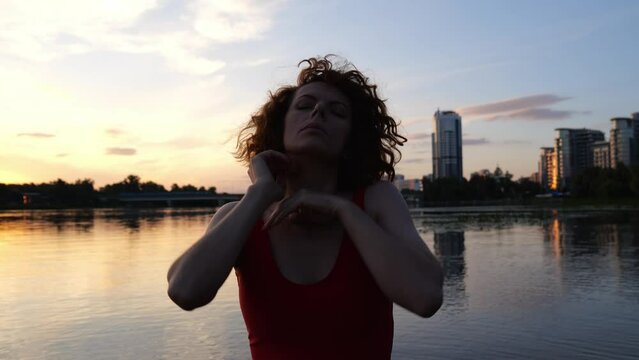 Portrait of a young redhead woman near dark water against the backdrop of a city in the distance. Slow motion woman in a bathing suit makes a movement with her hands in the evening