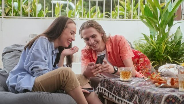 Medium shot of man showing meme picture to friend when hanging out together in outdoor cafe