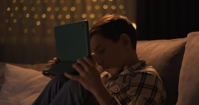 Tired kid falling asleep with gadjet in hands sitting on sofa at night. Sleepy kid sit on couch with tablet device