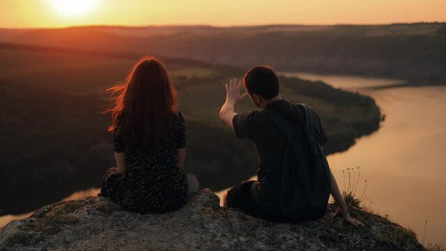 Couple man and woman looking at river view at sunset, romantic vacation during travel. Couple watching sunset, talking on cliff edge above river in nature, back view.