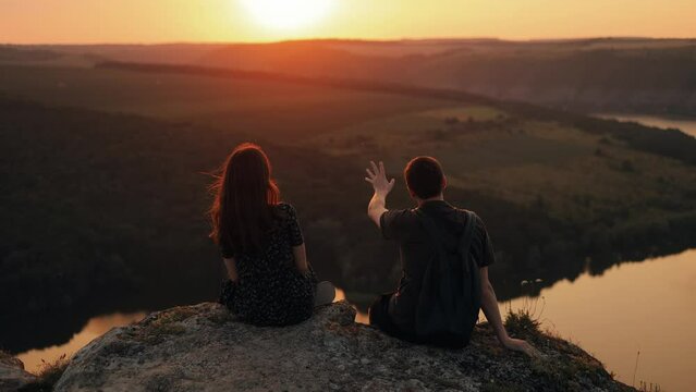Couple man and woman looking at river view at sunset, romantic vacation during travel. Meeting of young people. Couple watching sunset, talking on cliff edge above river in nature, back view.